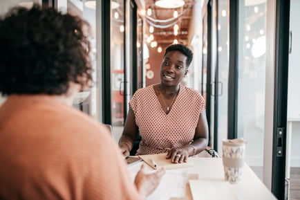 Two women, sitting and talking at a desk over coffee